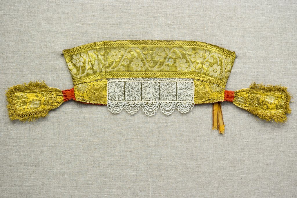 Девичий головной убор - повязка, вышитая золотыми цветами и листьями на зеленовато-золотом фоне. По центру — ажурный узор из белого бисера, собранного в виде снежинок, под ним в несколько рядов белые бисерные полукружья. По бокам — завязки золотистого цве