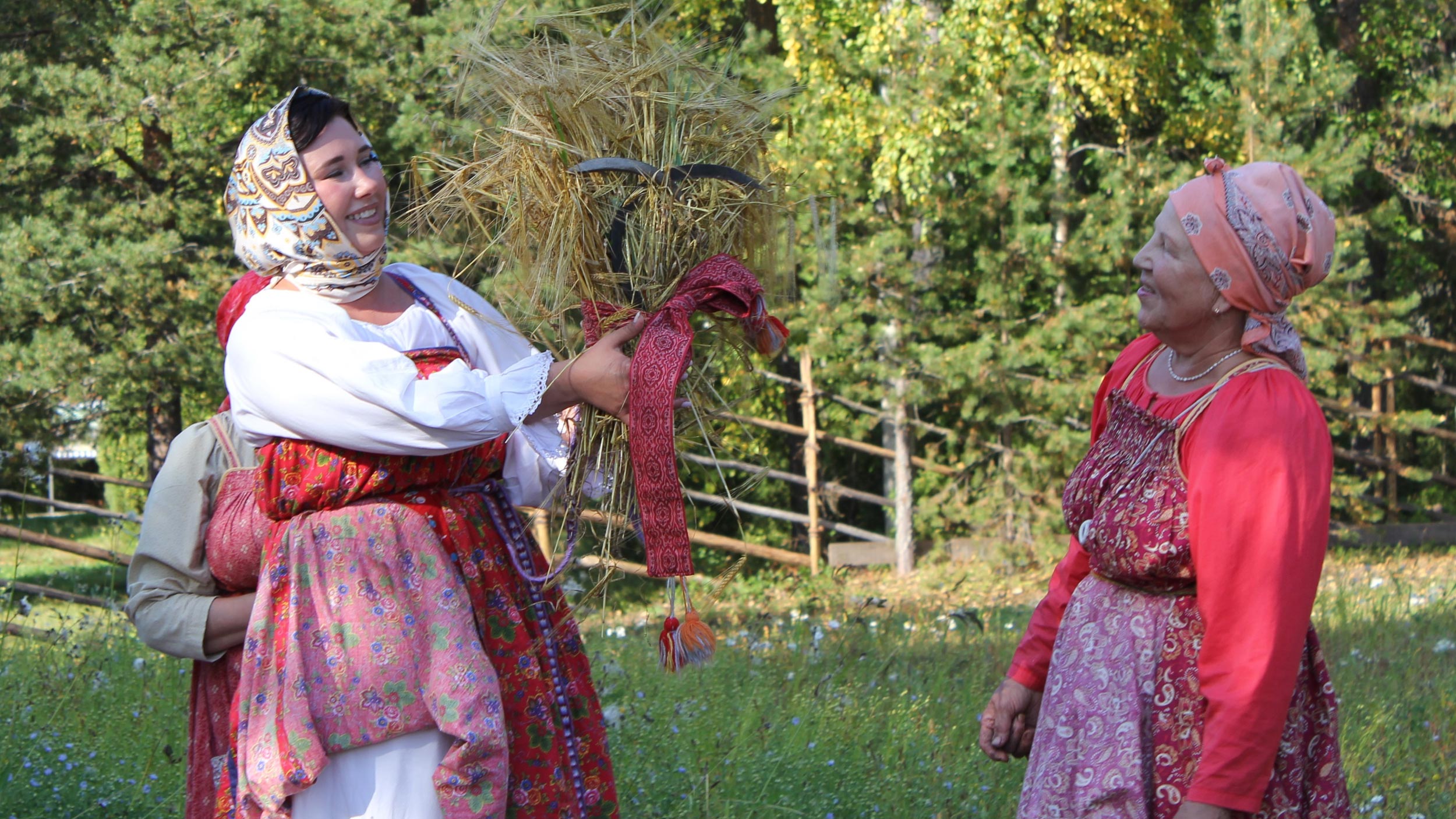 На фоне природы и деревьев стоят две женщины в традиционных нарядах. Одна из них держит в левой руке сноп, а в правой серп. Вторая ей улыбается, слегка наклонив голову. Это момент с этнокультурной фестиваля «Борода», посвящëнного сбору урожая