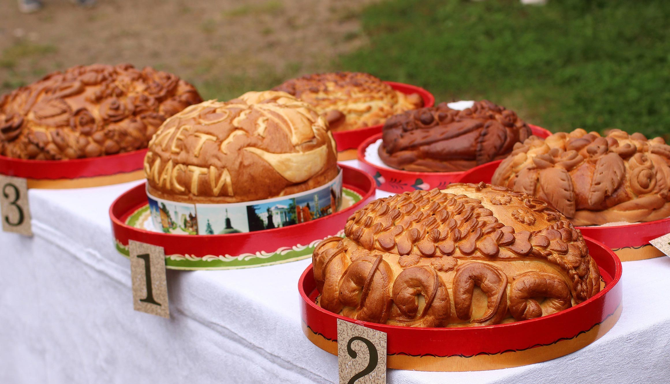 Множество художественно оформленных караваев, расположенных на столе. Каждый хлеб имеет уникальный декор, надписи и узоры. Внизу - подпись 