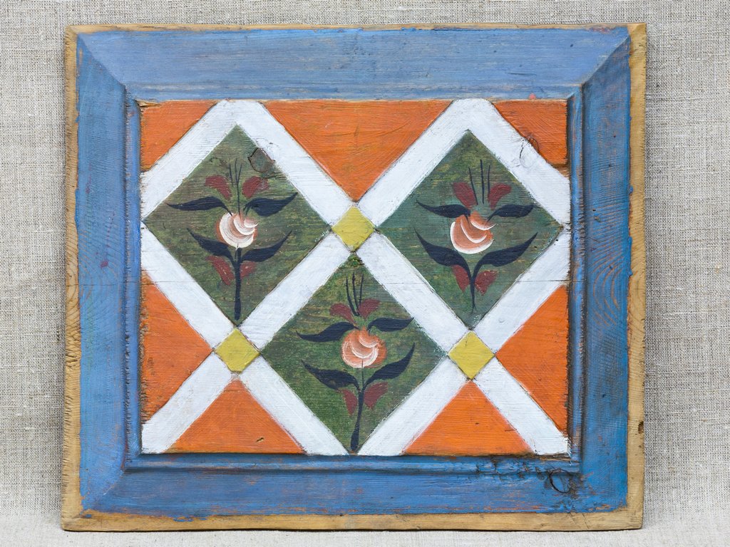 Деревянная прямоугольная панель с ярким орнаментом. Фон разделен на ромбообразные секции оранжевого, зеленого и белого цветов, украшенные стилизованными цветами. Рама выполнена в голубом цвете