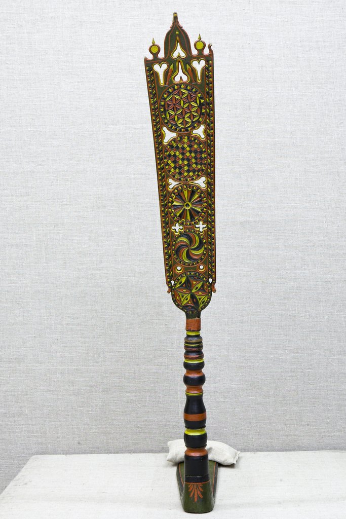 Деревянная расписная прялка с ажурной узорчатой верхушкой и резным орнаментом, стоящая вертикально; ручка гладкая, украшена цветными кольцами, основание плоское, темное