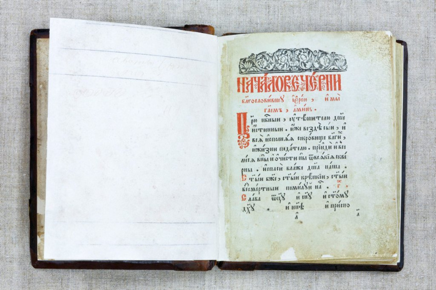Разворот старинной толстой книги на светлой ткани. Слева белый чистый лист, справа страница с кириллическим текстом, выполненным старинным шрифтом, первая буква крупная, красного цвета. Вверху страницы декоративная чёрная заставка с орнаментом.