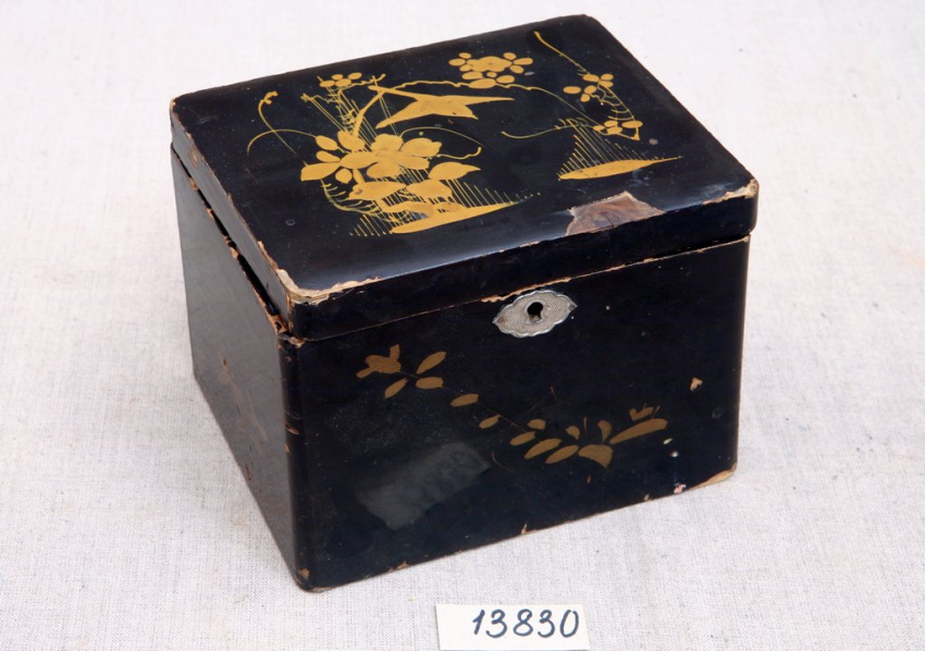 Прямоугольная чёрная лакированная шкатулка с потëртостями. На крышке и стенках  растительный орнамент золотистого цвета, на передней части — скважина для замка