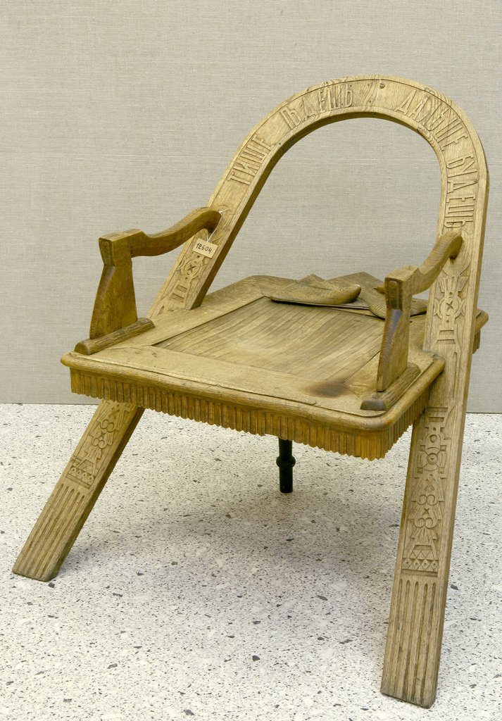 Старинное деревянное кресло светлого цвета с изогнутой спинкой и невысокими подлокотниками. Спинка и ножки украшены резьбой с орнаментом и надписью.