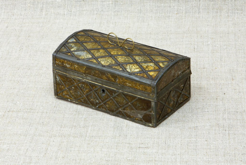 Прямоугольная шкатулка с выпуклой крышкой. Поверхность сделана из полупрозрачных жёлто-коричневых пластин, обрамлëнных металлической решёткой с ромбами. На крышке тонкая золотистая ручка-петелька, спереди маленькое отверстие для замка.