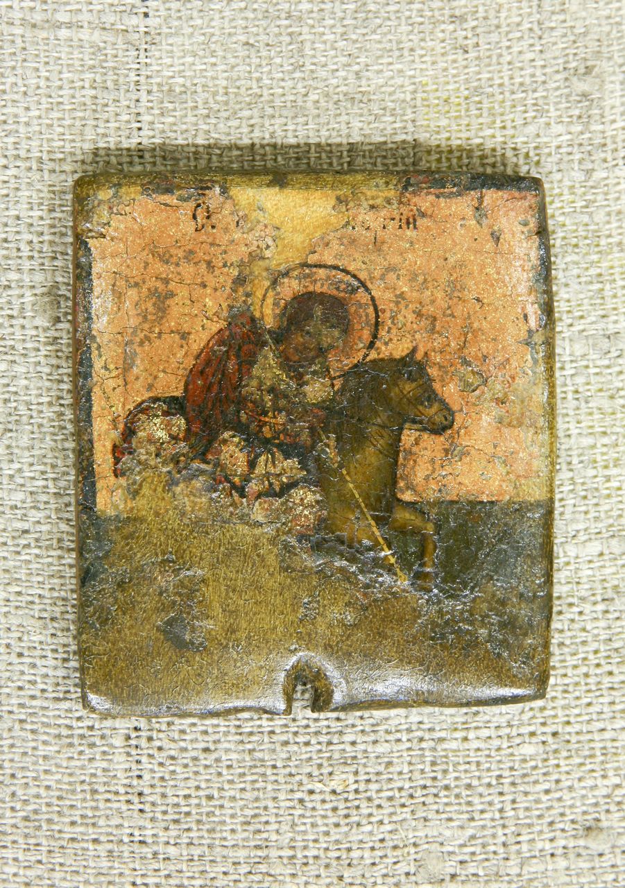 Небольшая икона с изношенной поверхностью. На ней изображëн святой в красной мантии на коричневой лошади, с ореолом над головой и копьём в руке. Фон оранжево-коричневый, часть иконы стëрта