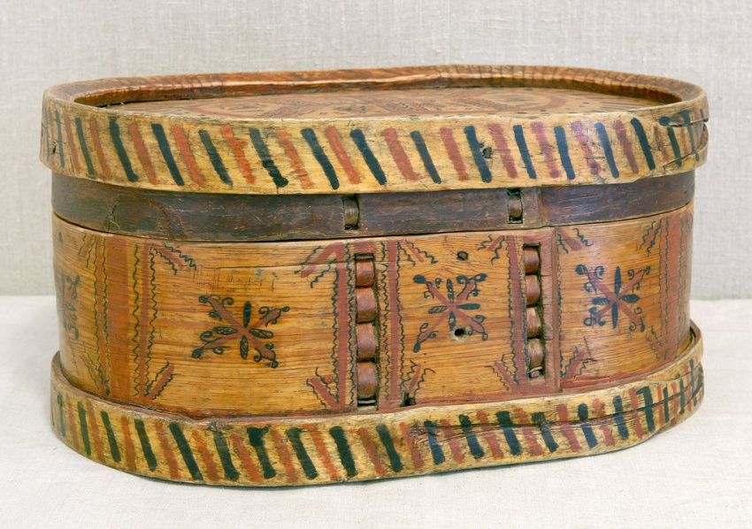 Овальная деревянная коробка с крышкой, украшенная узорами. На боковой поверхности — геометрические и растительные мотивы, по краям - чёрно-красный орнамент в виде чередующихся диагональных полос