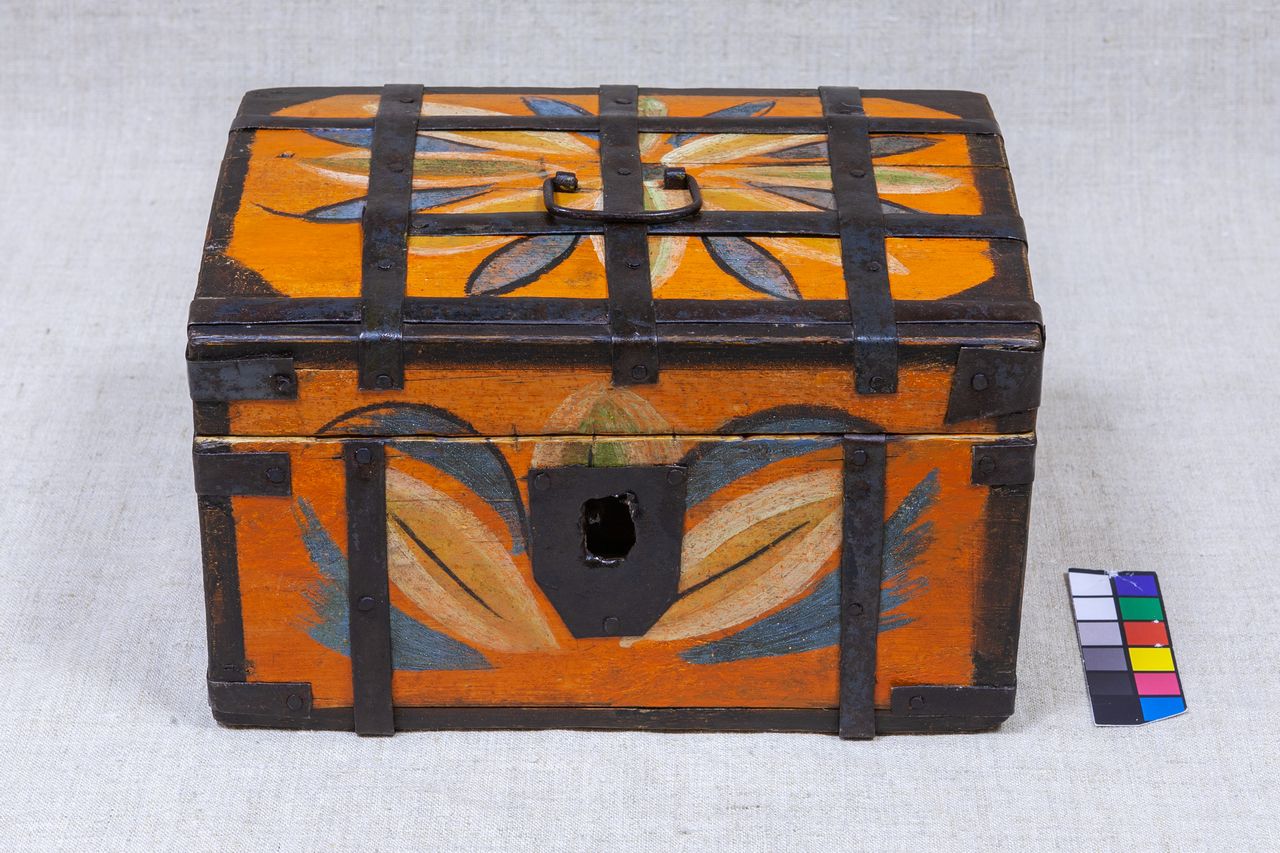 Деревянный сундук с железными накладками и ручкой сверху. Сундук ярко-оранжевый, украшен крупными разноцветными цветочными узорами на крышке и передней стороне