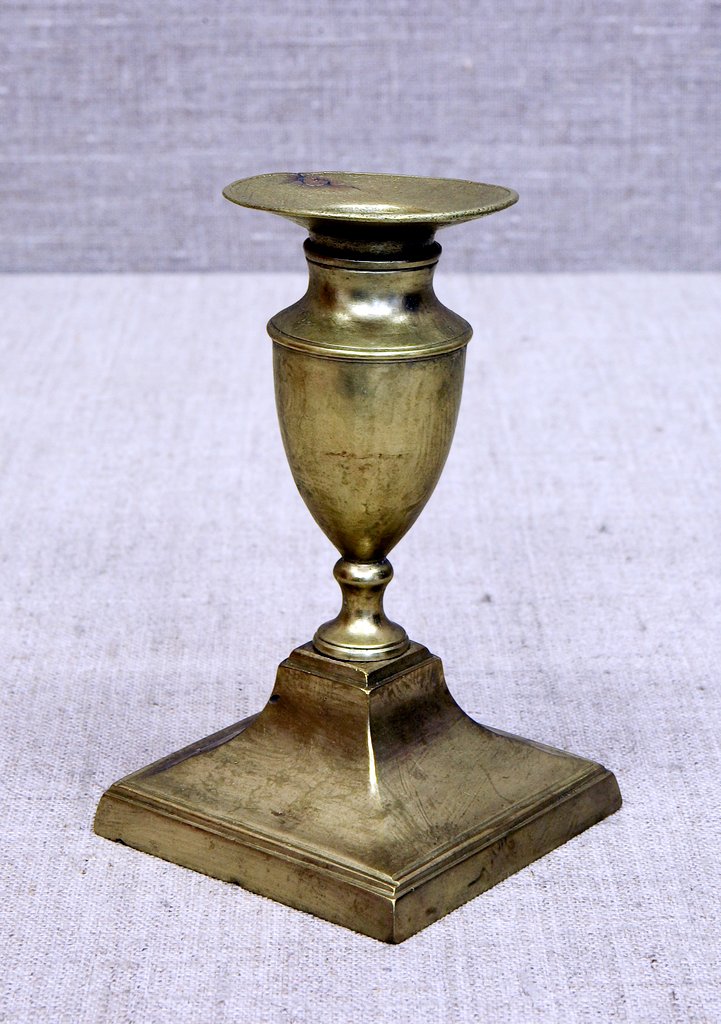 Металлический подсвечник золотистого цвета на широком квадратном основании с узкой ножкой и овальным бокалом‑держателем. Сверху расположено круглое плоское место для свечи.