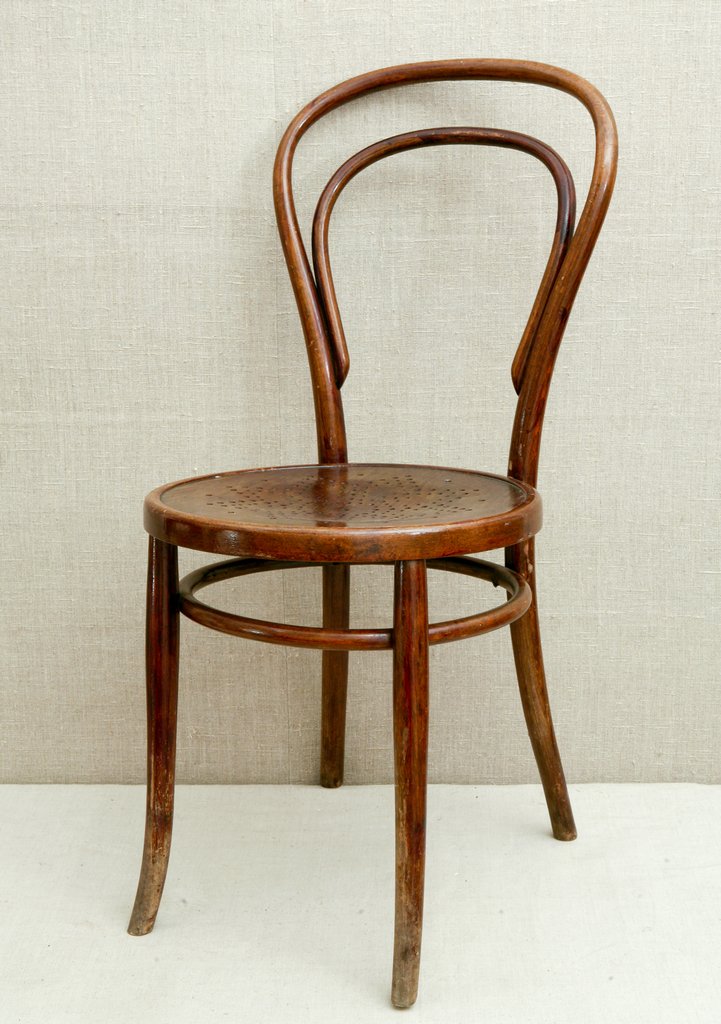 Деревянный стул с изогнутой спинкой и круглыми сиденьем стоит на светлом фоне. У стула четыре изогнутые ножки и сквозные отверстия на поверхности сиденья.