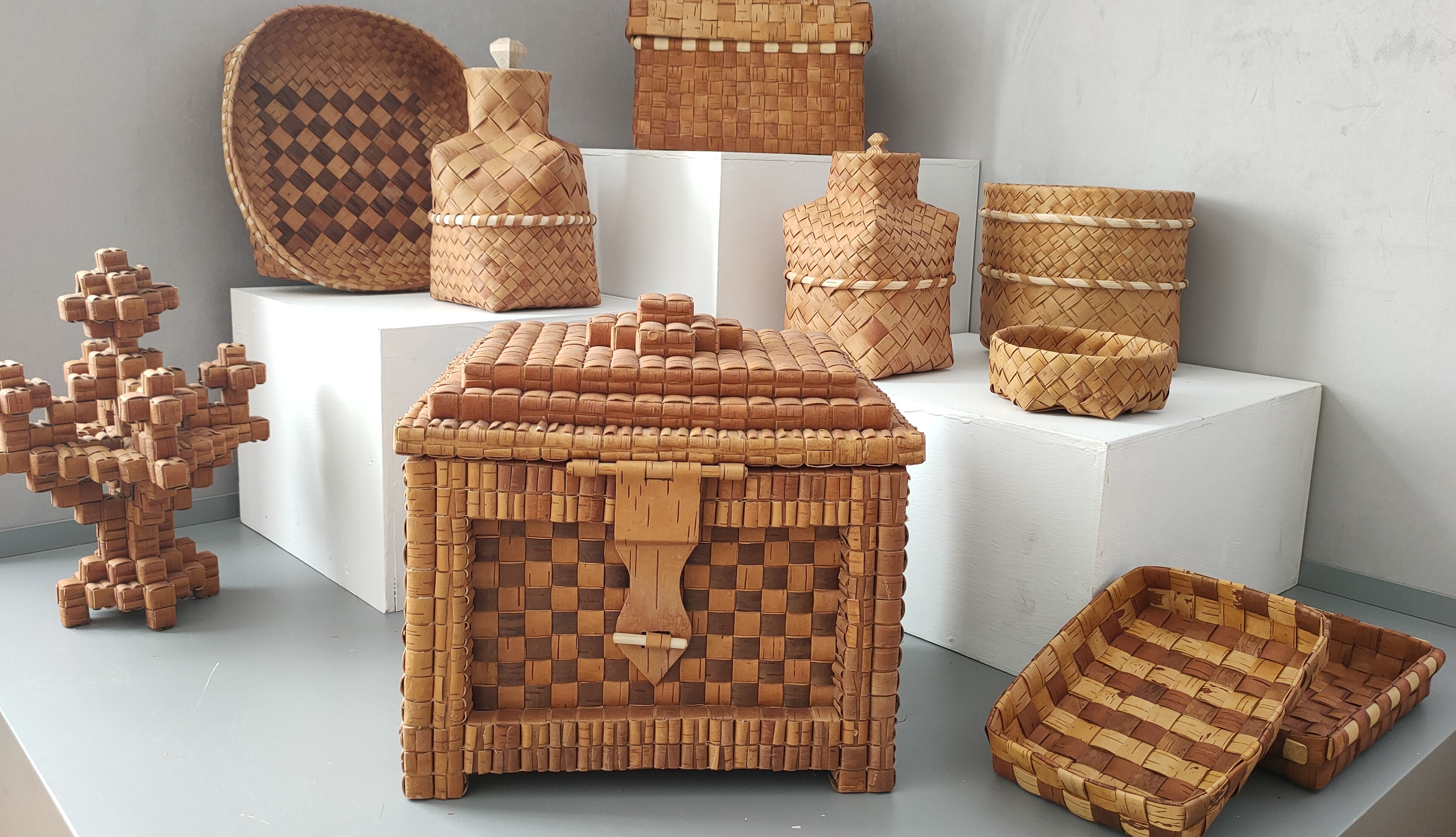 На изображении представлены изделия народного ремесла из плетеных материалов. В центре - плетеная коробка с крышкой, рядом расположены различные контейнеры и украшения, такие как плетеное дерево. На фоне видна надпись, связанная с темой выставки