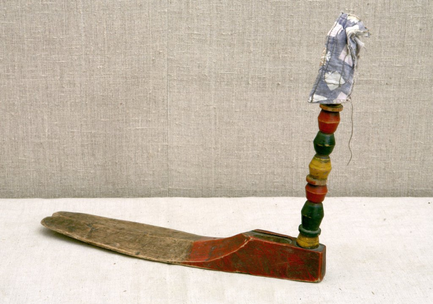 Деревянный предмет, похожий на гладкую лопатку с приподнятым задним краем, окрашенным красным. Сзади вертикально закреплён столбик из круглых разноцветных бусин, сверху на них надет маленький лоскуток ткани как чепчик.