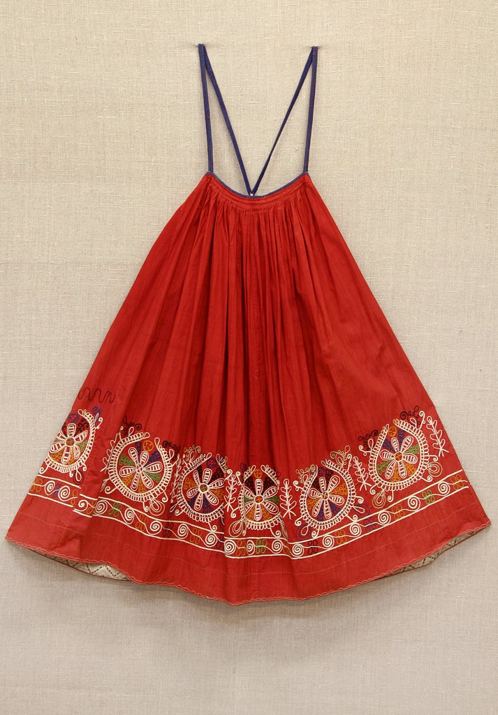 Красный сарафан с тонкими тёмно‑синими бретелями. Низ украшен цветочной вышивкой и орнаментальной каймой; верх собран частыми мелкими складками у талии