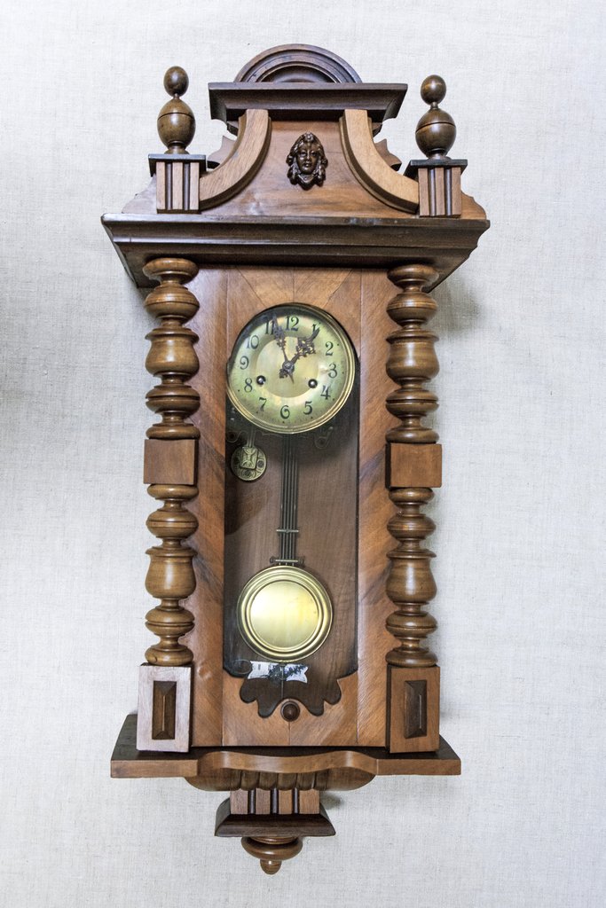 Настенные часы в резном деревянном корпусе в классическом стиле. Вверху фигурный декор с шариками и маской, в центре под стеклом круглый циферблат и длинный маятник с круглым грузом.