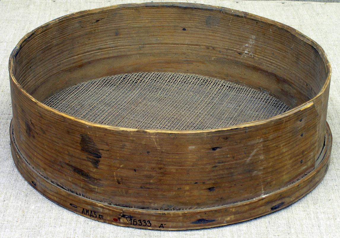 Круглое деревянное сито с высокими стенками и сетчатым дном. Материал имеет потёртости и тёмные пятна. Сито используется для просеивания муки