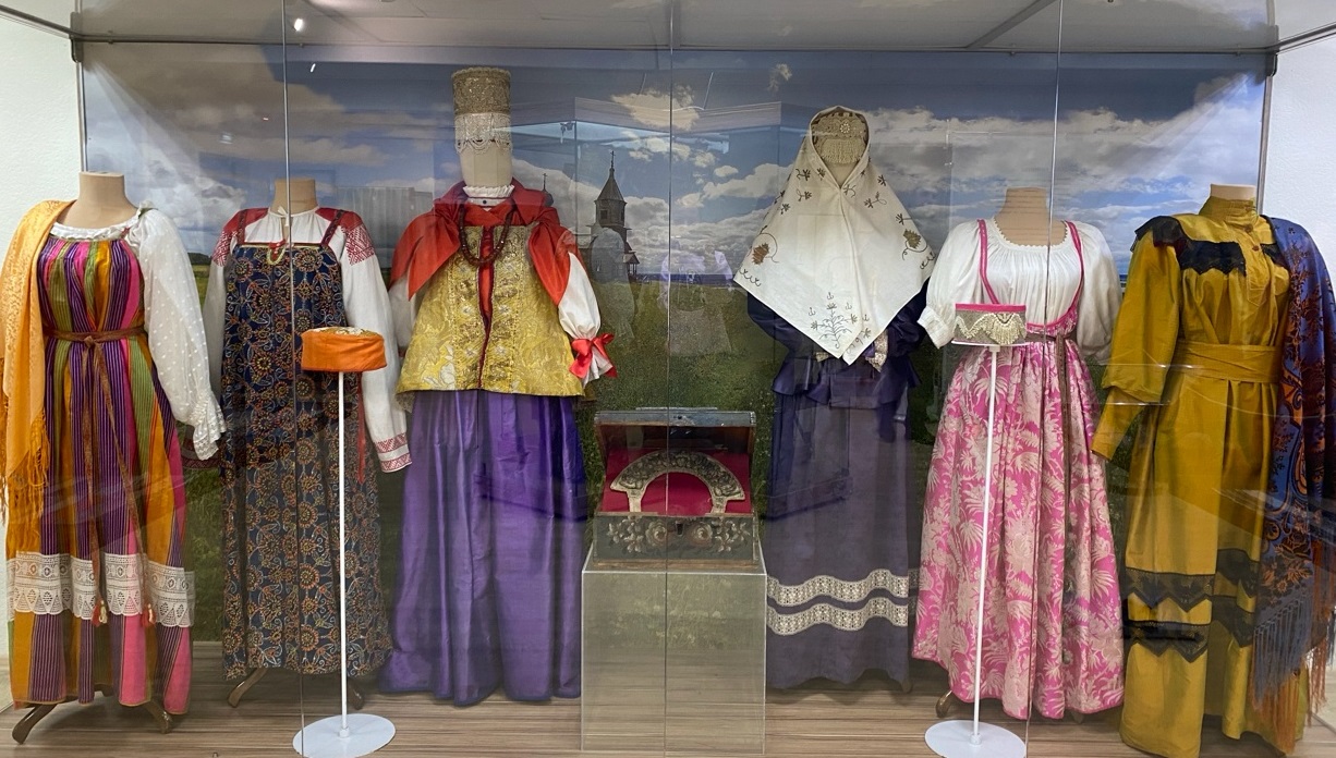 Народные костюмы, выставленные в витрине музея. Костюмы разнообразны по цвету и стилю: от ярких и веселых до более сдержанных и традиционных. Надпись анонсирует выставку под названием «Будто пава»