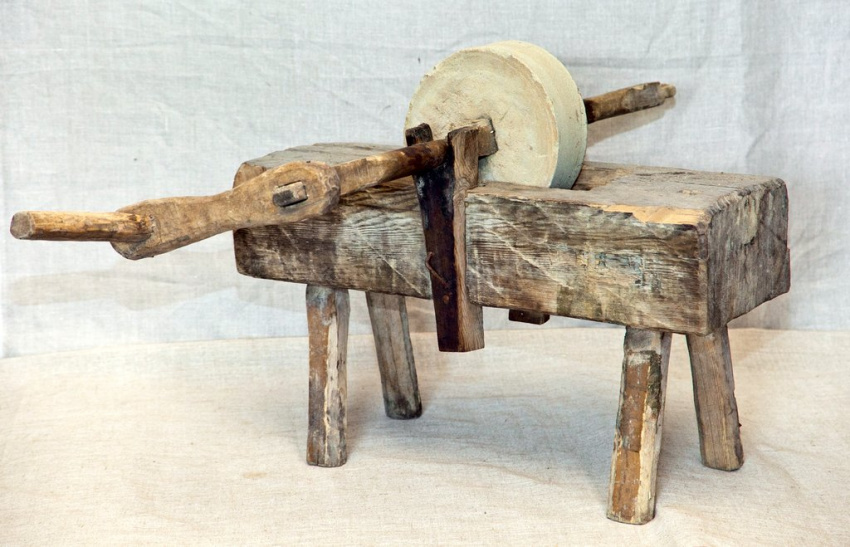 Старинный точильный станок: основа в виде деревянного четырехгранного бруска на четырёх ножках, в верхней части которого посередине точильный круг из камня, сбоку длинная деревянная рукоять для вращения круга вручную.