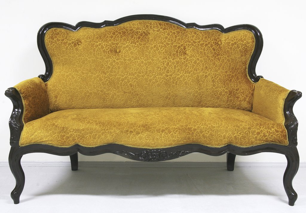 Старинный трёхместный диван с изогнутой резной деревянной рамой тёмного цвета. Обивка дивана выполнена из бархатистой ткани жёлто-горчичного оттенка с узорчатым рисунком. Диван стоит на четырёх изогнутых ножках и выглядит массивно и элегантно.
