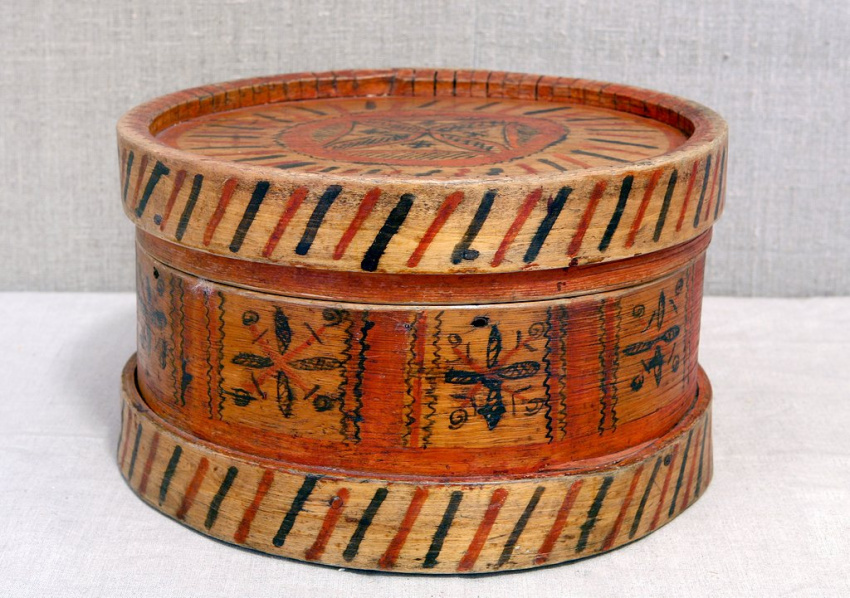 Круглая деревянная коробка с крышкой. По краям полосатый орнамент чёрного и красного цветов. На боках и крышке — геометрические узоры и цветы, выполненные в красно-коричневых цветах