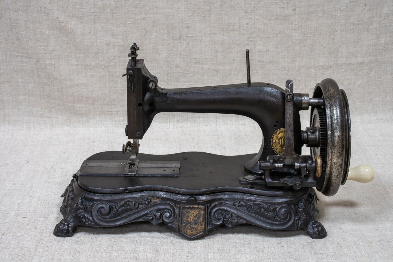 Старая механическая швейная машина на массивной металлической резной подставке, преобладают темные цвета. Машина выполнена из металла, с крупным маховым колесом справа