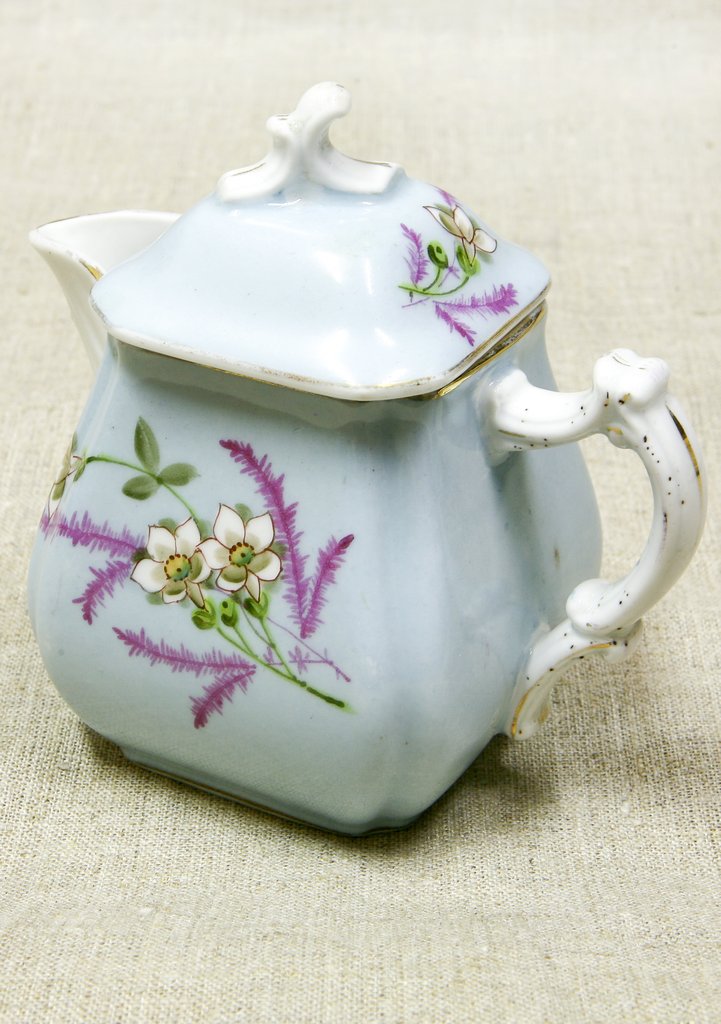 Четырёхгранный фарфоровый кувшин белого цвета с крышкой, изогнутой ручкой и носиком. Стороны украшены рисунком из белых цветов и тонких фиолетовых веточек, узор крышки повторяет основной.