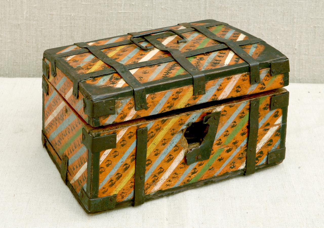 Старый деревянный сундук с металлическими уголками и ремнями. Корпус окрашен яркими полосами оранжевого, желтого и голубого цвета. На передней части отверстие для замка. Крышка сундука приоткрыта