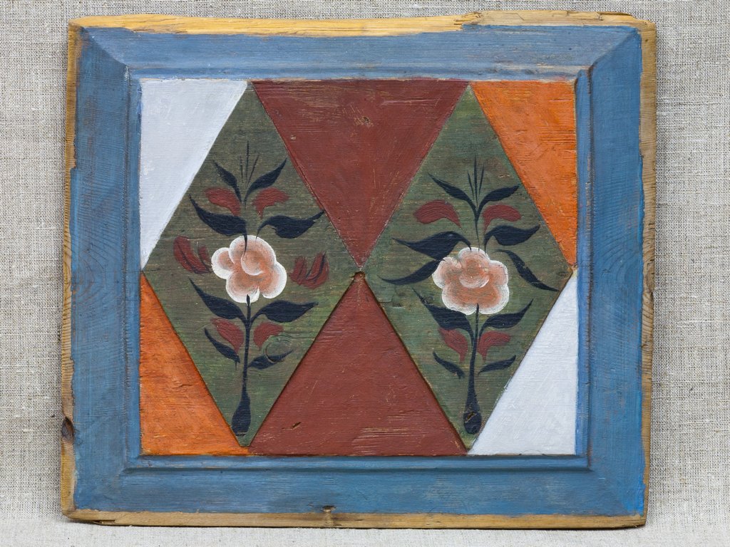 Декоративная деревянная панель прямоугольной формы. Периметр обведëн голубыми цветом, в центре разноцветные ромбы и треугольники. В каждом ромбе изображения цветов с лепестками розового цвета. Фон в теплых и холодных тонах, создавая яркий контраст