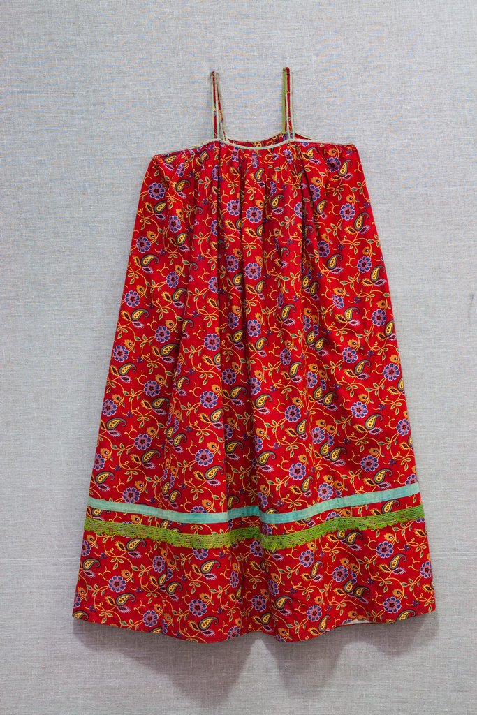 Ситцевый сарафан на тонких бретелях. Ярко-красный фон покрыт мелким рисунком из фиолетовых цветов и жёлтых завитков. Верхняя часть собрана в крупные не частые складки, по низу две горизонтальные декоративные тесьмы зелёного и бирюзового цвета