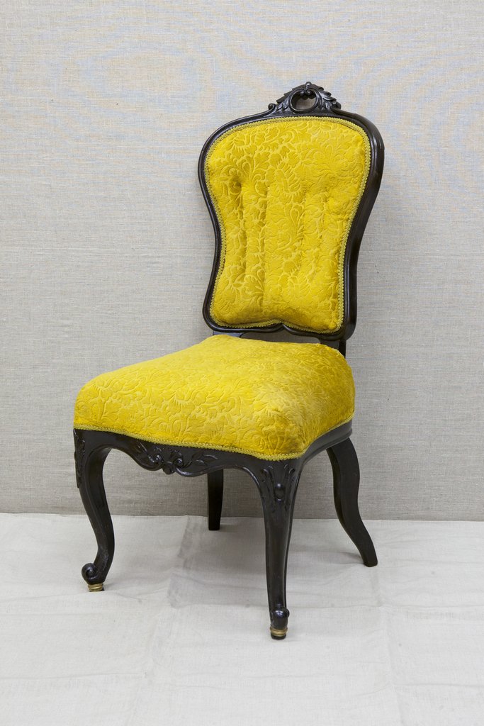 Стул без подлокотников. Спинка и сиденье обиты яркой бархатистой тканью жёлтого цвета. Каркас стула выполнен из тёмного резного дерева: на верхней части спинки изящный орнамент. Ножки стула изогнуты и придают ему элегантный внешний вид.