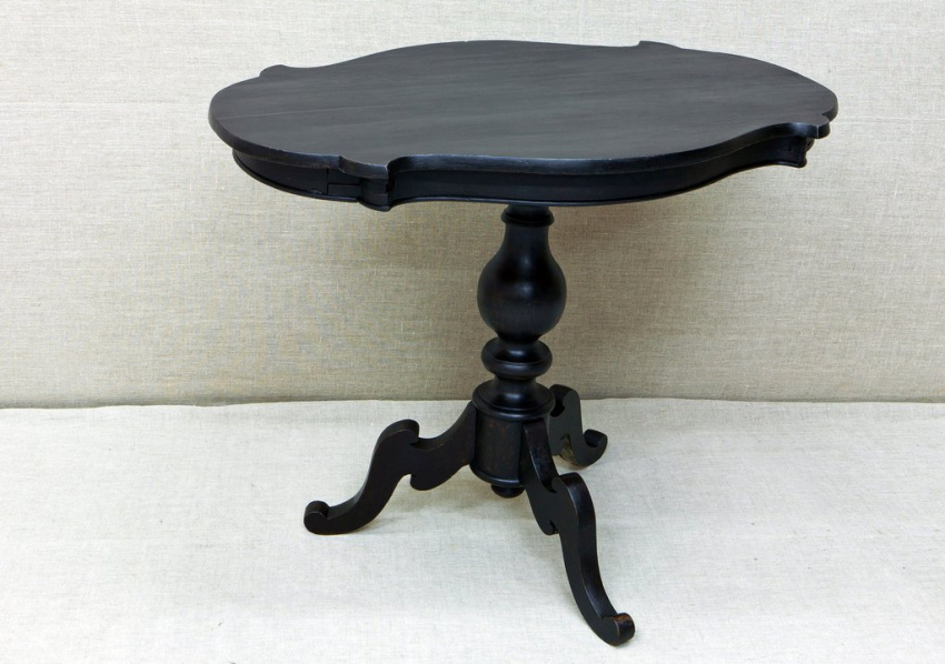 Невысокий круглый столик из тёмного дерева. Столешница овальная с волнистыми краями, поверхность пустая. Ножка фигурная, плавно сужается кверху. Внизу три изогнутые опоры с завитками.