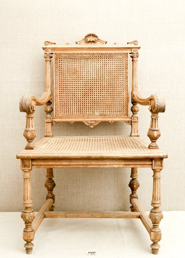 Деревянный стул с высокой спинкой и массивными изогнутыми подлокотниками, украшенный сложной резьбой: завитки, шарики, растительные орнаменты. Спинка и сиденье с плетёными вставками. Стул светлого матового оттенка толстые ножки с резьбой и перекладинами.
