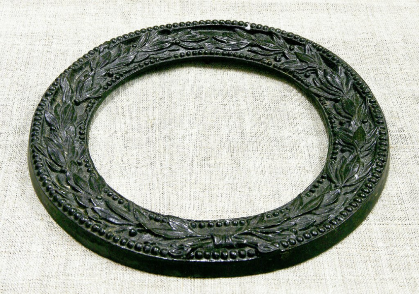 Литая из чугуна рама, выполненная в виде кольца. По внешнему и внутреннему краям декорирована рельефными шарами, между которыми расположен рельефный венок из лавровых веток, скрепленный внизу по центру бантом.