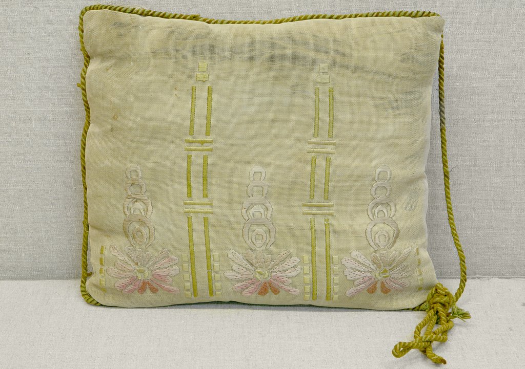 Светлая квадратная подушка с вышивкой в виде вертикальных линий и узоров, а также цветком в нижней части. Края подушки украшены зеленым закрученным шнуром, угол завершён кисточкой из того же шнура.