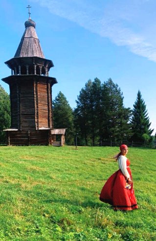 На фотографии летний пейзаж. На переднем плане девушка в народном костюме. На заднем – деревянная колокольня