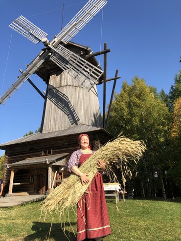 На фотографии изображена девушка в народном костюме со большим снопом. На заднем плане деревянная ветряная мельница