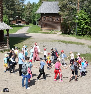 На фотографии девушка в народном костюме водит хоровод с детьми на площади. На заднем плане деревянный дом