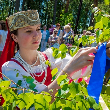 На фотографии изображена девушка в народном костюме, повязывающая ленточку на берёзовую ветвь
